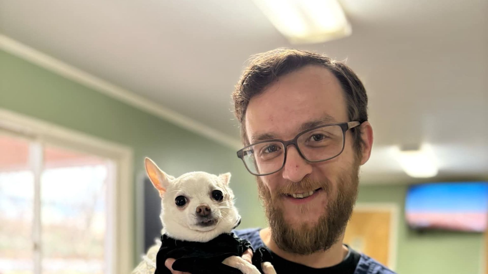 A man wearing glasses is holding a small white dog in his arms